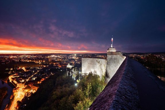 Historische Festungsanlage über einer beleuchteten Stadt bei Sonnenuntergang und violettem Abendhimmel
